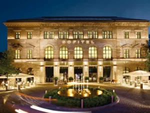 The illuminated 19th-century neoclassical facade of Sofitel Munich Bayerpost, formerly the Royal Bavarian Post Office, featuring grand arched windows and a modern fountain at the entrance.