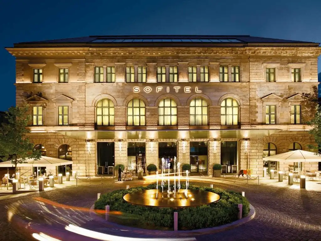 The illuminated 19th-century neoclassical facade of Sofitel Munich Bayerpost, formerly the Royal Bavarian Post Office, featuring grand arched windows and a modern fountain at the entrance.