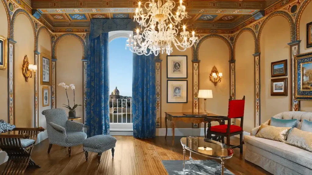 A lavishly decorated suite at The St. Regis Florence, featuring ornate wall frescoes, a crystal chandelier, and a balcony window offering a direct view of the Arno River and the San Frediano in Cestello dome.