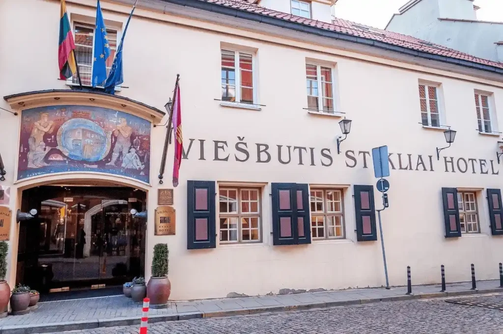The exterior of the Stikliai Hotel Vilnius, featuring the historic arched entrance topped by a traditional artisan-themed mural and the original wood-shuttered windows of the 16th-century complex.