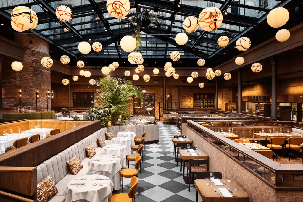The light-filled Root Restaurant at Telegraphenamt Berlin, located in a former courtyard under a massive 1910 industrial glass roof, featuring original red brickwork and contemporary globe lighting.