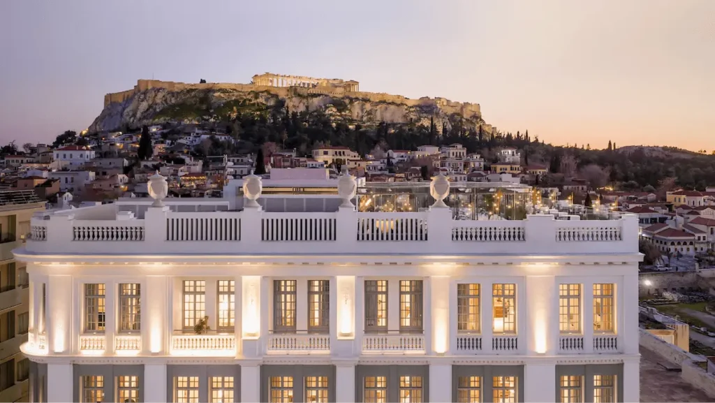 An exterior view of The Dolli at Athens, a luxury hotel housed in a 1920s neoclassical mansion designed by architect Andreas Kriezis, featuring its illuminated white facade and rooftop terrace with a direct, unobstructed view of the Parthenon and the Acropolis at sunset.
