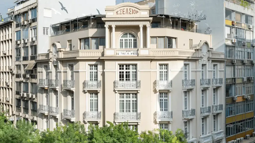 An exterior view of The Excelsior Thessaloniki, a luxury boutique hotel housed in a landmark 1924 neoclassical building designed by architects Pleyber and Fernandez, featuring its ornate balconies and iconic "EXCELSIOR" lettering on the rooftop.