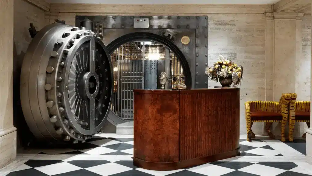 The iconic The Ned London bank vault entrance, featuring a massive 25-tonne circular stainless steel door from the building's 1920s history as the Midland Bank headquarters, set against a checkerboard floor.