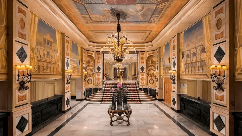 The grand neoclassical lobby of The Palace Madrid, featuring marble columns, ornate ceiling frescoes, and a restored 1912 glass palm-tree chandelier, reflecting the hotel’s €90 million restoration completed in March 2025.