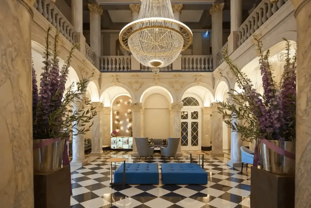 The spectacular six-story central atrium of The Ritz-Carlton Hotel de la Paix Geneva, featuring a checkered floor, marble columns, and a grand crystal chandelier.
