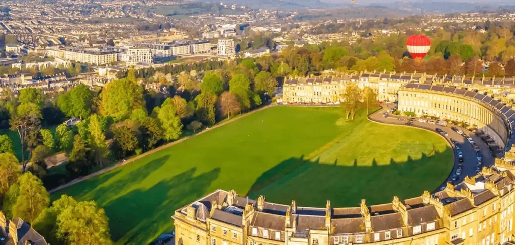 An aerial perspective of The Royal Crescent Hotel & Spa in Bath, showcasing the sweeping honey-colored Georgian architecture of the 1775 crescent, the expansive Royal Victoria Park lawn, and a hot air balloon rising over the historic city.