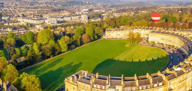 An aerial perspective of The Royal Crescent Hotel & Spa in Bath, showcasing the sweeping honey-colored Georgian architecture of the 1775 crescent, the expansive Royal Victoria Park lawn, and a hot air balloon rising over the historic city.