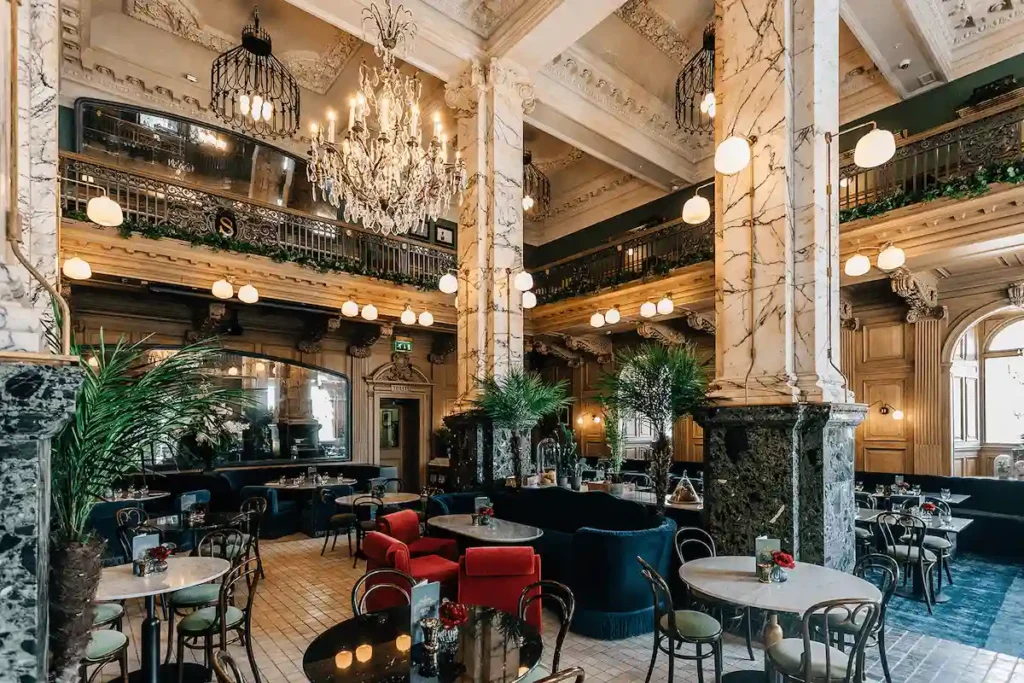 The double-height Grand Café at The Scotsman Hotel in Edinburgh, featuring original massive marble pillars, ornate Edwardian wood carvings, and crystal chandeliers in the building's former newspaper advertising hall.