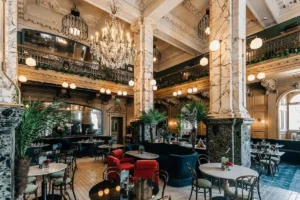 The double-height Grand Café at The Scotsman Hotel in Edinburgh, featuring original massive marble pillars, ornate Edwardian wood carvings, and crystal chandeliers in the building's former newspaper advertising hall.