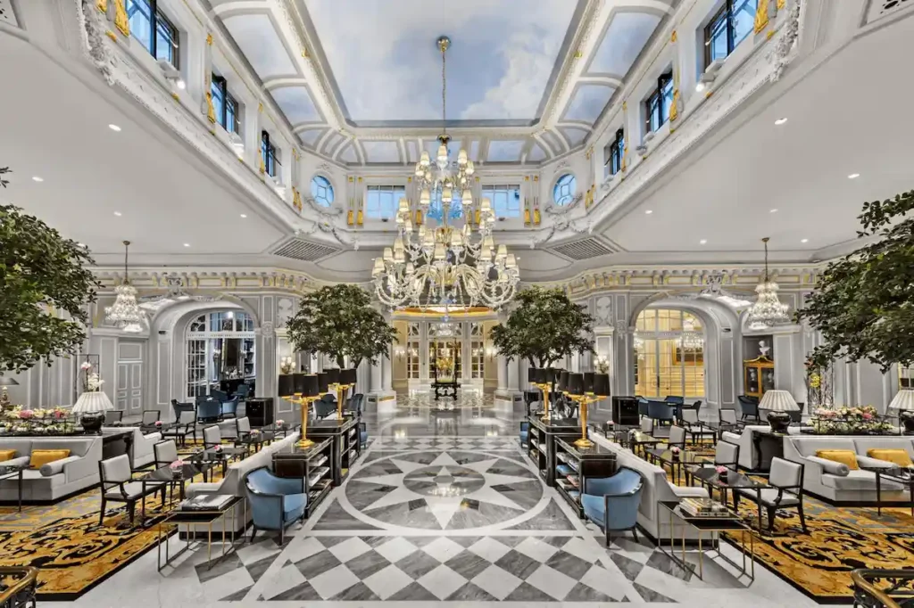 The opulent Grand Hall of The St. Regis Rome, a historic 1894 palazzo, featuring a soaring skylight with a trompe l'oeil cloud ceiling, massive crystal chandeliers, and a checkered marble floor.