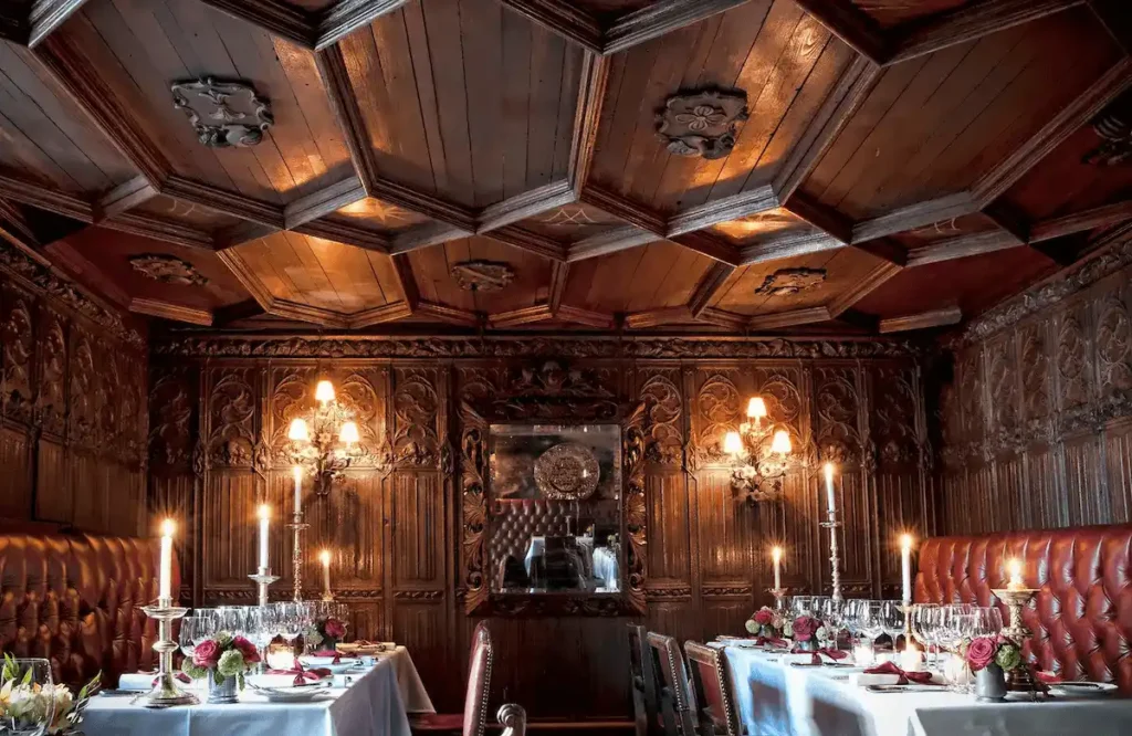 The candlelit dining room of The Witchery by the Castle in Edinburgh, featuring dark oak-paneled walls with intricate carvings, a magnificent 17th-century painted timber ceiling, and red leather banquettes at tables set for fine dining.