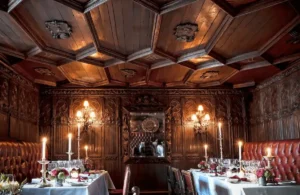 The candlelit dining room of The Witchery by the Castle in Edinburgh, featuring dark oak-paneled walls with intricate carvings, a magnificent 17th-century painted timber ceiling, and red leather banquettes at tables set for fine dining.