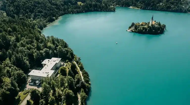 An aerial view of the modernist white stone Vila Bled estate nestled in a dense green forest on the shore of Lake Bled, with the iconic island and pilgrimage church in the turquoise water nearby.