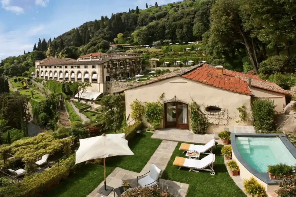 A wide-angle view of the Limonaia Villa at Villa San Michele Florence, featuring a private plunge pool, manicured lawn, and sun loungers, with the historic monastery building and terraced Fiesole gardens in the background.
