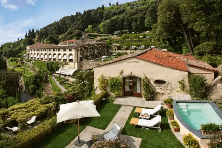 A wide-angle view of the Limonaia Villa at Villa San Michele Florence, featuring a private plunge pool, manicured lawn, and sun loungers, with the historic monastery building and terraced Fiesole gardens in the background.