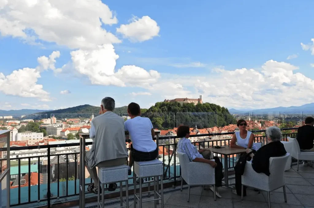 3 Day Itinerary in Slovenia travel guide image showing the open-air rooftop terrace bar of the Nebotičnik Skyscraper, with guests seated at tables looking out over a panoramic view of Ljubljana's old town and Ljubljana Castle on its green hill under a blue sky with white clouds.