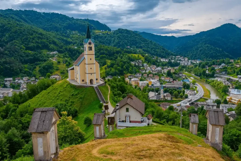 5 Day Itinerary in Slovenia alternative travel guide image showcasing a panoramic, high-angle view of the town of Idrija, highlighting the prominent yellow Church of St. Anthony (Cerkev sv. Antona Padovanskega) sitting on top of a lush green hill with the Idrijca River valley and forested mountains in the background.