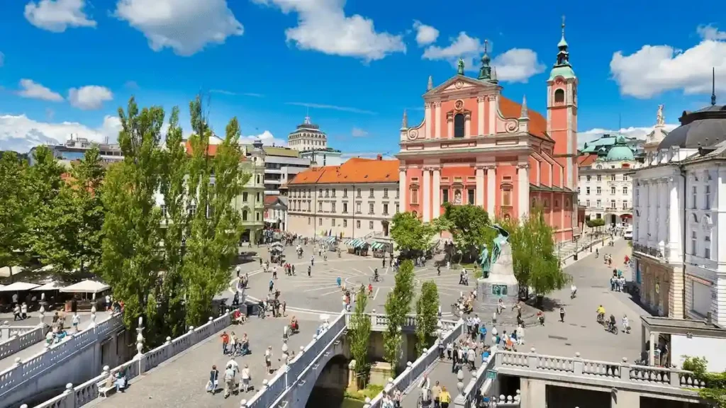 7 Day Itinerary in Slovenia guide image featuring an overhead view of Prešeren Square, the pink Franciscan Church of the Annunciation, and the iconic Triple Bridge spanning the Ljubljana River on a sunny day with travelers walking around.