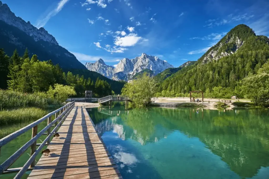 Best time to visit Slovenia travel guide image featuring a person mountain biking across a rustic wooden boardwalk on the crystal-clear, emerald waters of Lake Jasna in Kranjska Gora, with lush green forests and the dramatic, snow-capped peaks of the Julian Alps reflecting perfectly on the water under a clear blue sky.