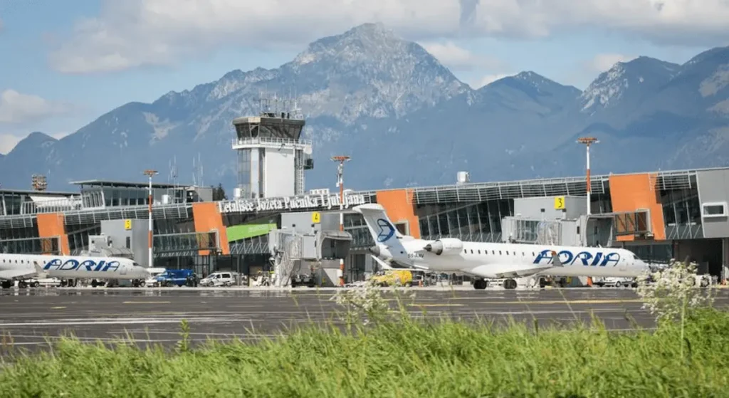 How to get from Ljubljana Airport to city center travel guide image showing the exterior tarmac and passenger terminal of Ljubljana Jože Pučnik Airport, featuring Adria Airways airplanes parked at the gates, the control tower, and the prominent Kamnik–Savinja Alps rising in the background.