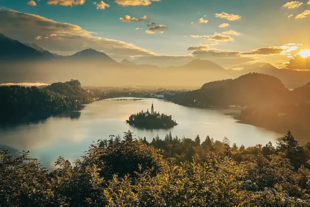 Slovenia honeymoon romantic guide image showing a golden sunrise over Lake Bled, with the iconic island church enveloped in a light morning mist, Bled Castle perched on the cliffside, and the majestic peaks of the Julian Alps rising in the background under a warm, sunlit sky.